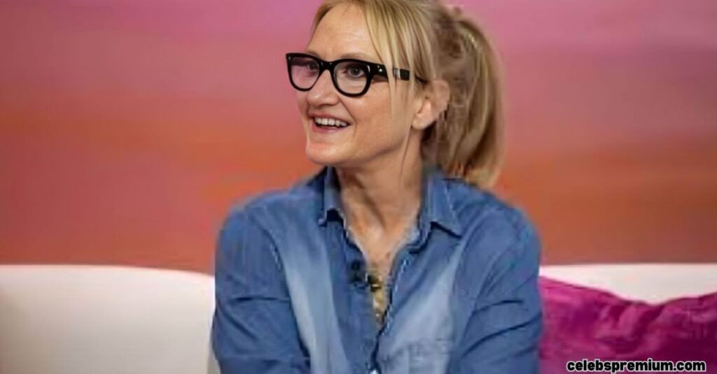 Mel Robbins, wearing black-framed glasses and a blue denim shirt, is smiling and looking to her left. Her blonde hair is pulled back in a ponytail. She appears to be seated on a white surface with a purple pillow behind her, against a blurred orange and pink background.