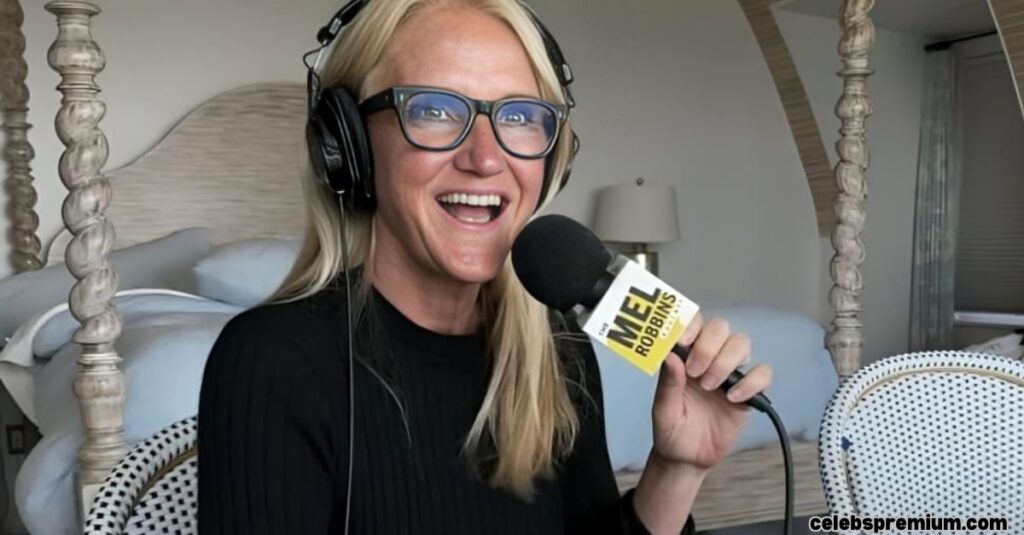 Mel Robbins, with long blonde hair and glasses, is enthusiastically speaking into a microphone with a "MEL ROBBINS" logo. She is wearing headphones and a black top, and appears to be recording a podcast or broadcast from a bedroom setting with a four-poster bed.