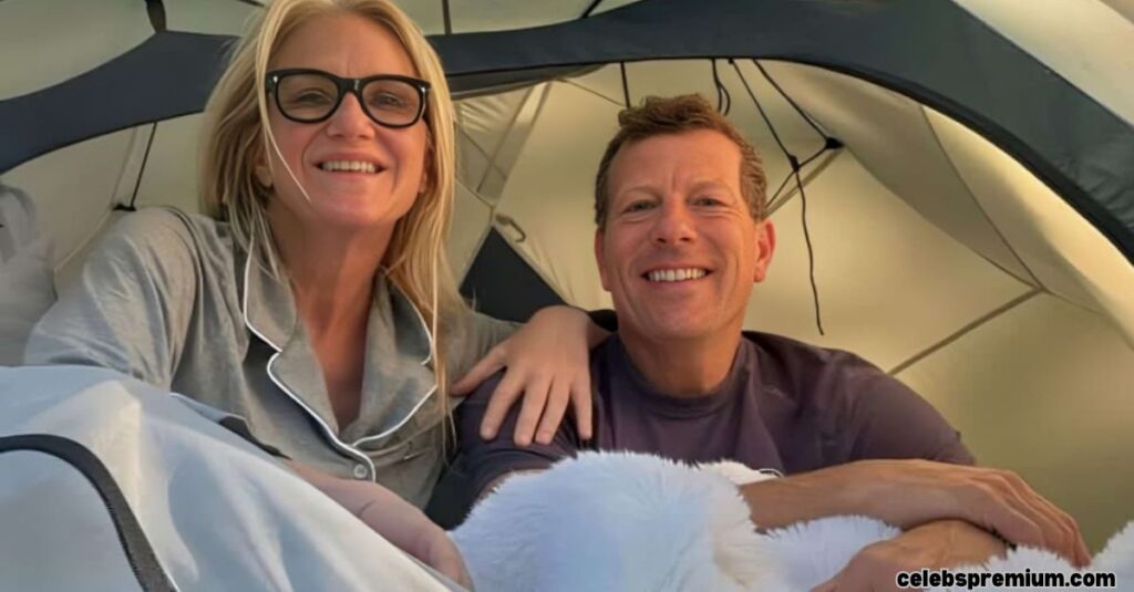 A photo of Mel Robbins and her husband inside a tent, both smiling at the camera. Mel is wearing pajamas and glasses, and her husband is in a casual shirt. They are under a white fluffy blanket, suggesting they are camping.