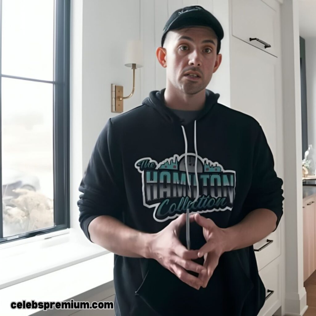 A photo of Steve Hamilton wearing a black cap and a black hoodie featuring "The Hamilton Collection" branding. He has his hands together and is looking to the side. The setting appears to be inside a modern home with a window and white cabinetry.