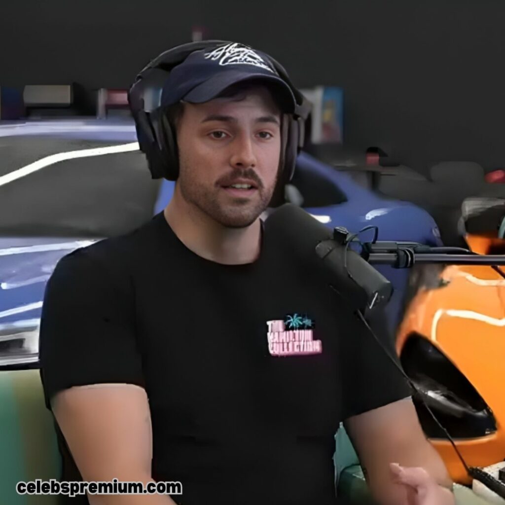 Steve Hamilton, wearing a black t-shirt and a dark baseball cap with white writing, is wearing black headphones and speaking into a microphone. He has a mustache and a goatee. The blurred background shows parts of luxury cars in blue and orange.