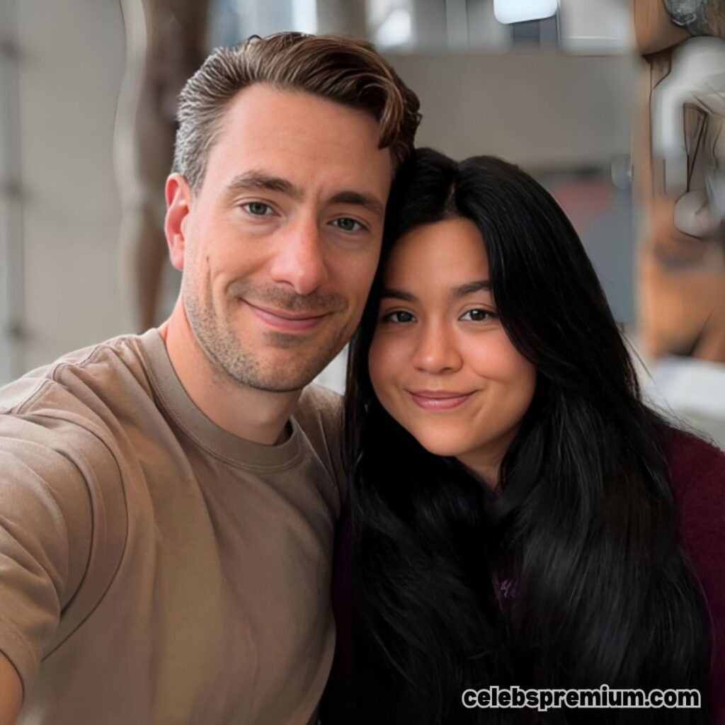 Andrew Wilkinson and his wife are posing for a selfie. Andrew has short brown hair and is wearing a dark brown coat over a white shirt. His wife has long dark hair and is wearing a maroon sweater.