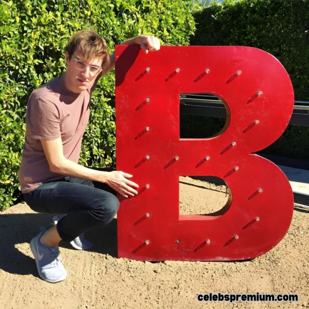 Andrew Wilkinson is crouching next to a large, red, illuminated letter "B" outdoors on a gravel surface. He is wearing glasses, a brown t-shirt, dark pants, and grey sneakers. Green foliage is visible in the background.