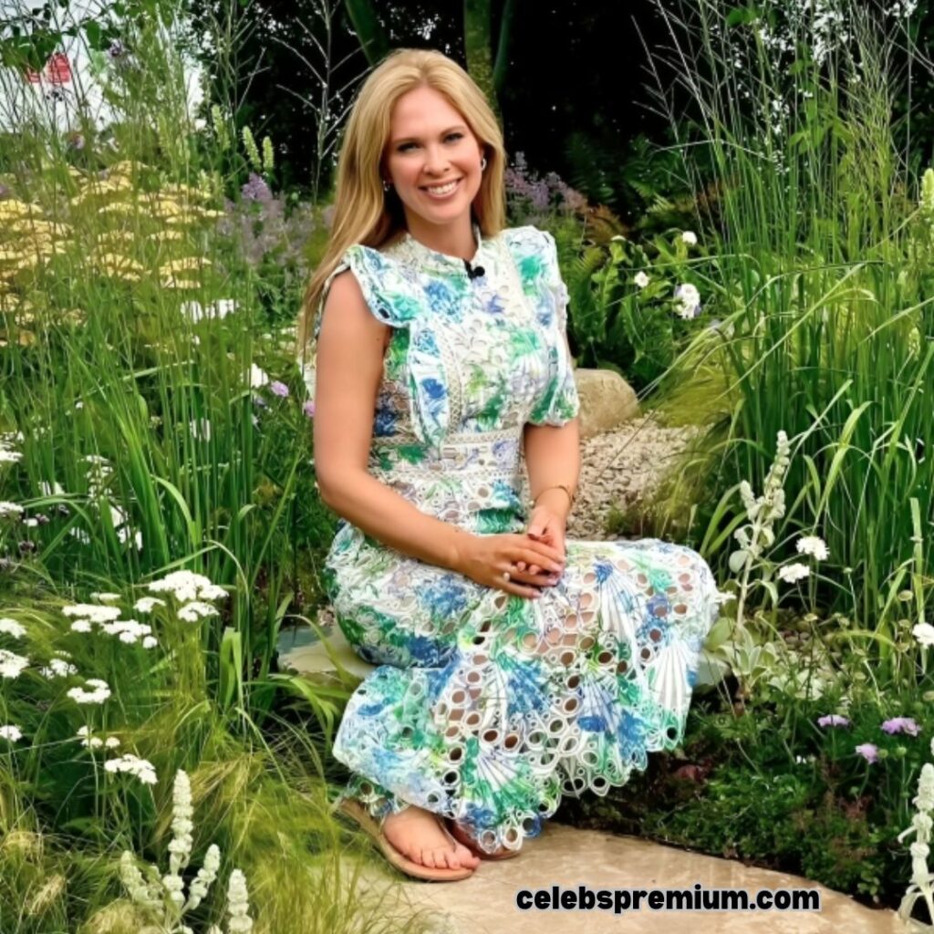 Jo Blythe is sitting on a stone in a lush garden, smiling at the camera. She is wearing a sleeveless midi dress with a white base and a blue and green floral pattern with eyelet details. The garden is filled with tall grasses, white wildflowers, and purple blooms.