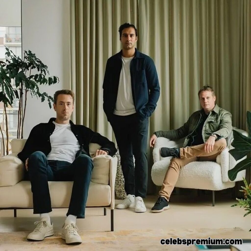 A photo shows Andrew Wilkinson sitting in a light-colored armchair, wearing a dark top and jeans. Standing behind him and slightly to the right is another man in a dark jacket and light-colored trousers. To the right, a third man is seated in a matching armchair, wearing a patterned jacket and tan pants. They are in a room with neutral tones, plants, and curtains.