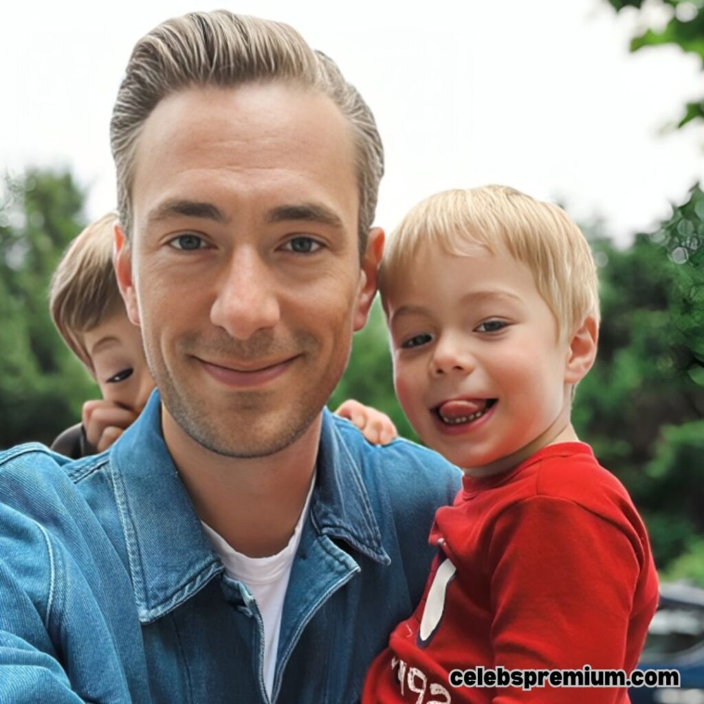 Andrew Wilkinson is taking a selfie with two young children. He is wearing a blue denim jacket. One child with blonde hair is in his arms, sticking out their tongue and smiling. Another child with brown hair is peeking from behind Andrew's shoulder. The background is blurry with greenery.