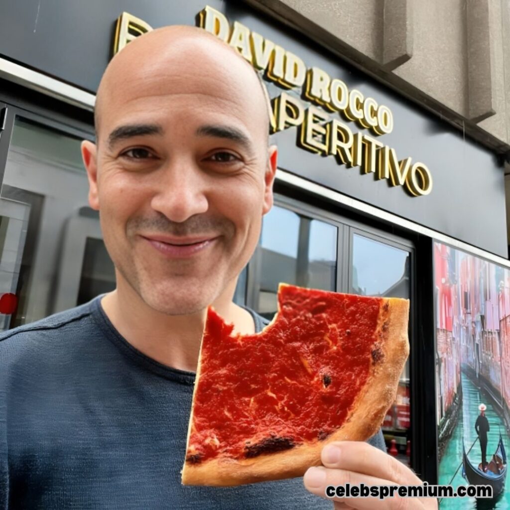 David Rocco holds a slice of red pizza with a bite taken out of it, smiling at the camera. Behind him is a shopfront with "DAVID ROCCO APERITIVO" written above the entrance.