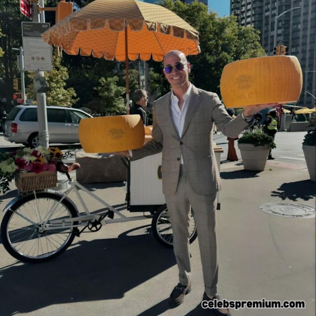 A photo of David Rocco in a tan suit and sunglasses, smiling and holding two large parmesan cheese wheels. He is standing near a bicycle cart with a yellow umbrella on a city street.