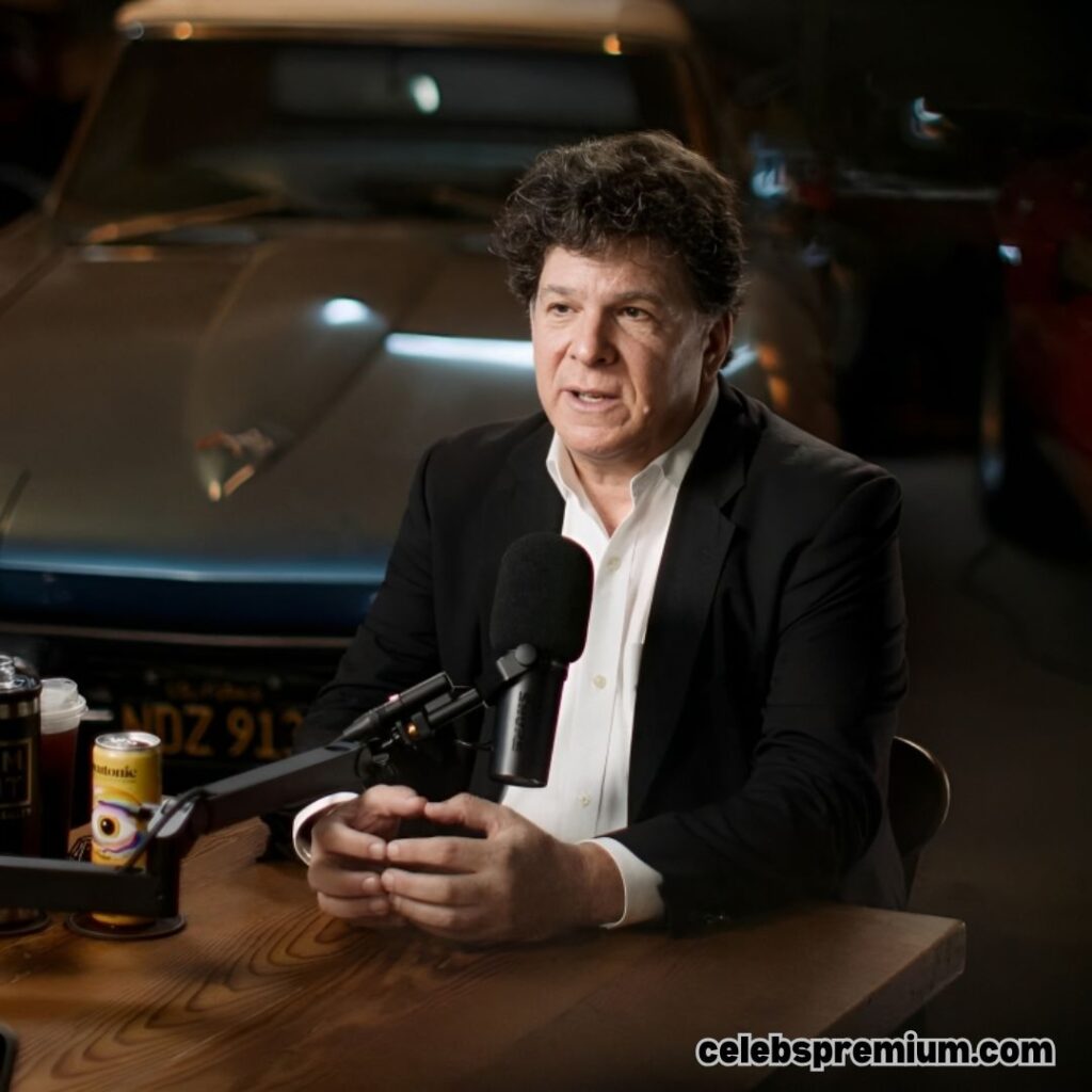 Eric Weinstein, wearing a black suit jacket over a white shirt, is speaking into a microphone. He is seated at a wooden table with his hands clasped. The blurred background shows classic cars.