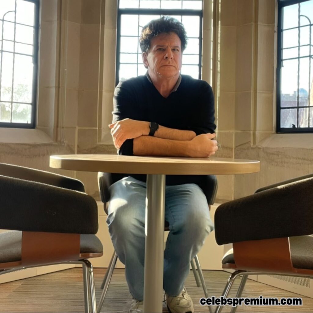 Eric Weinstein is seated at a round table inside a building with large windows. He is wearing a black long-sleeved shirt and light blue jeans. He has a serious expression on his face.