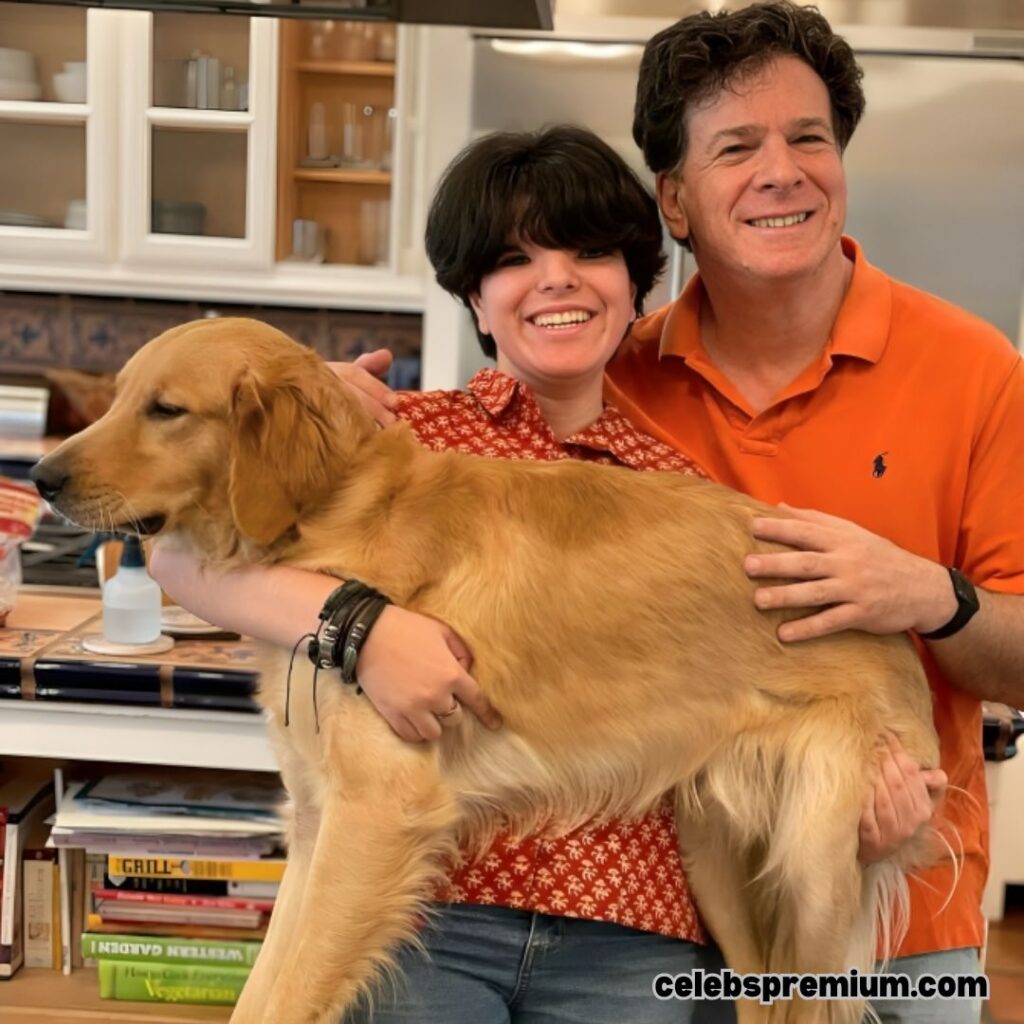 Eric Weinstein, wearing an orange polo shirt, is smiling with his arm around his son with dark hair wearing a red patterned shirt, who is holding a golden retriever dog. They are indoors, possibly in a kitchen, with cookbooks visible in the foreground.