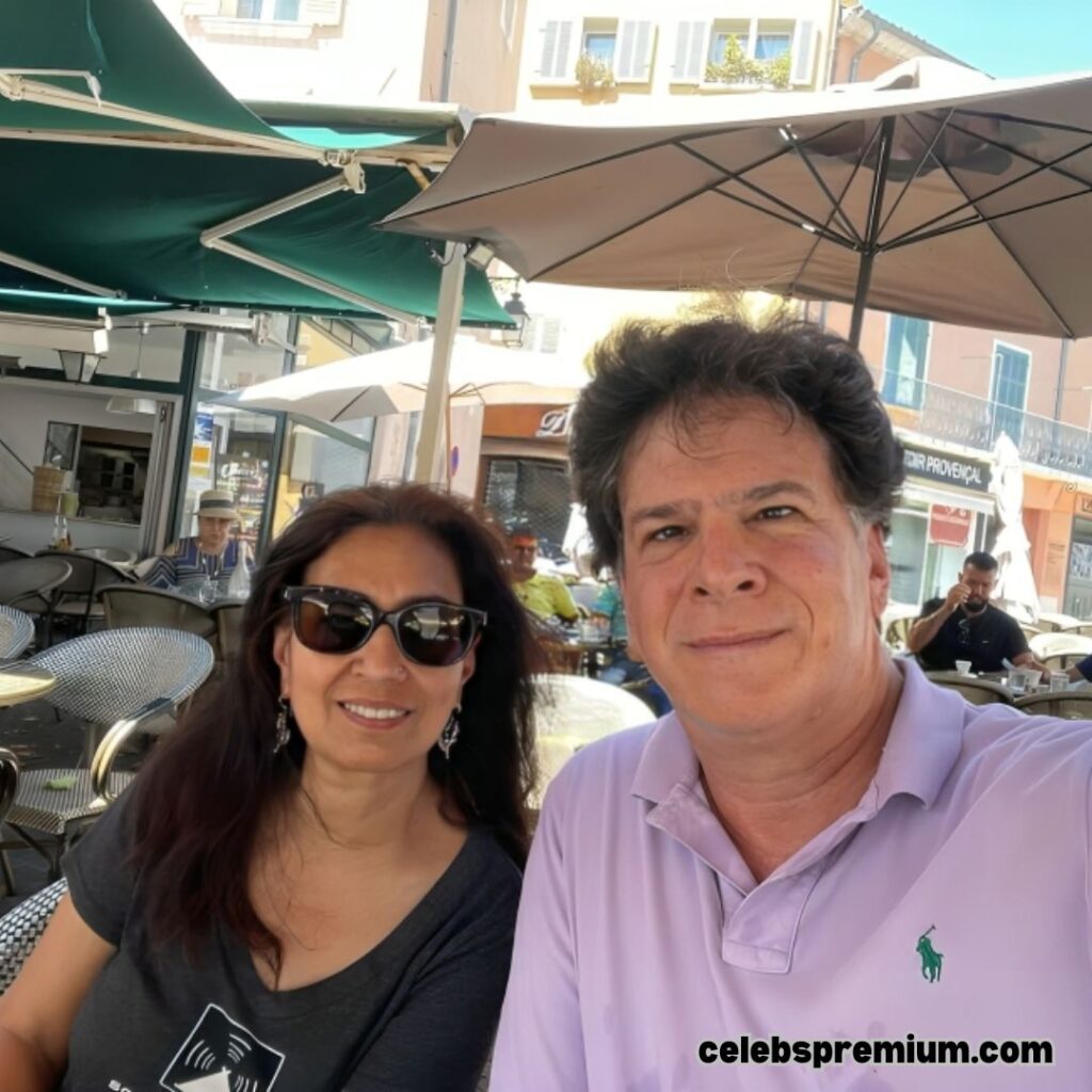 Eric Weinstein is taking a selfie with a woman with long dark hair and sunglasses at an outdoor cafe. Eric is wearing a light purple polo shirt. They are seated under umbrellas at a table with other cafe patrons and buildings in the background.