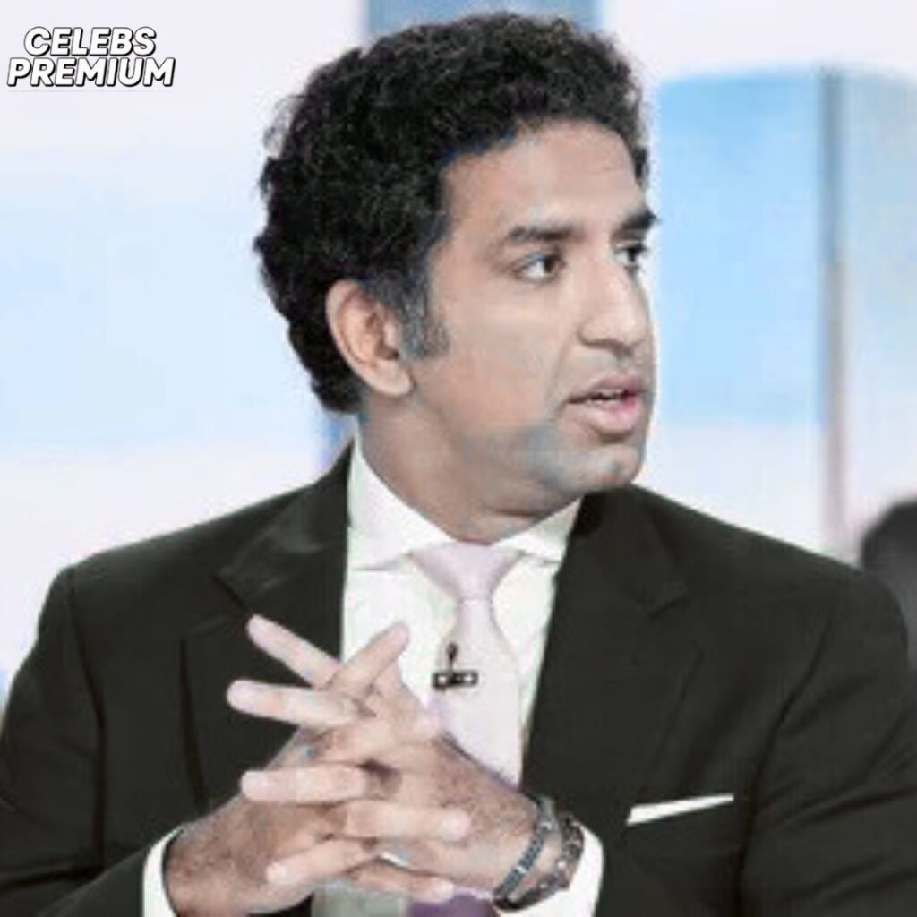 Omeed Malik, with dark curly hair, is wearing a black suit and a light pink tie. He has his hands clasped and is looking slightly to his right.