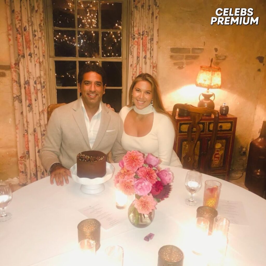 A photo of Omeed Malik and his wife at a dinner table. Omeed is wearing a light-colored suit jacket, and his wife is wearing a white dress. A chocolate cake and pink flowers decorate the table.