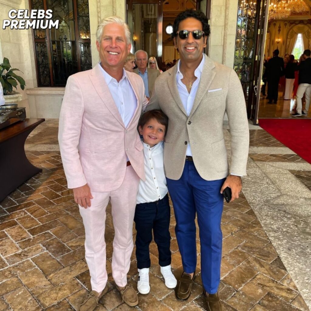 Omeed Malik, wearing a beige blazer and blue pants, stands with his arm around a young boy and a man in a pink suit. The boy is smiling, and the adults are looking at the camera. They are indoors, possibly at a formal event.