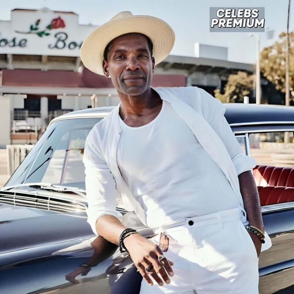 Ray Campbell, wearing a white shirt and pants with a light-colored jacket, leans against the hood of a classic car. He is also wearing a light-colored hat.