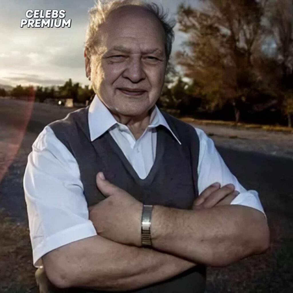 A photo of Ronald Wayne standing outside with his arms crossed. He is wearing a white shirt and a grey vest. The background shows a road and a sky with clouds.