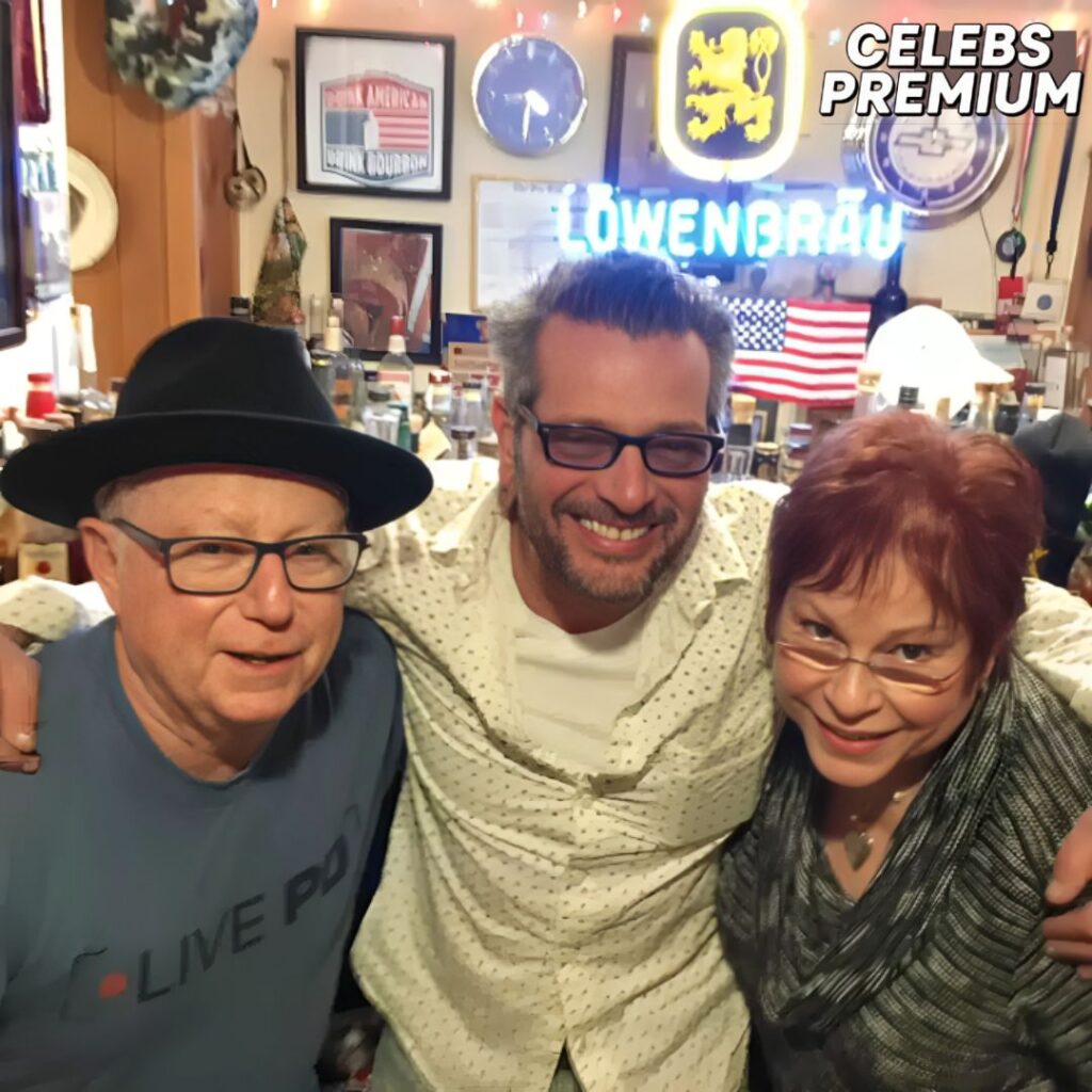 Scott LoBaido, wearing glasses and a light-colored patterned shirt, is in the center, smiling with his arms around two other people. The person on the left is wearing a black hat and glasses and a blue t-shirt. The person on the right has red hair and is wearing glasses and a patterned scarf. They are indoors in a cluttered room with various signs and decorations on the walls, including an American flag and a Löwenbräu neon sign.