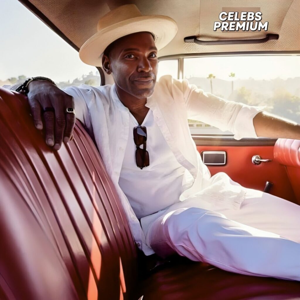 Ray Campbell, wearing a light-colored hat and white clothing, is sitting in the passenger seat of a car with red leather interior. Sunglasses are hanging from his shirt. He is looking towards the camera.