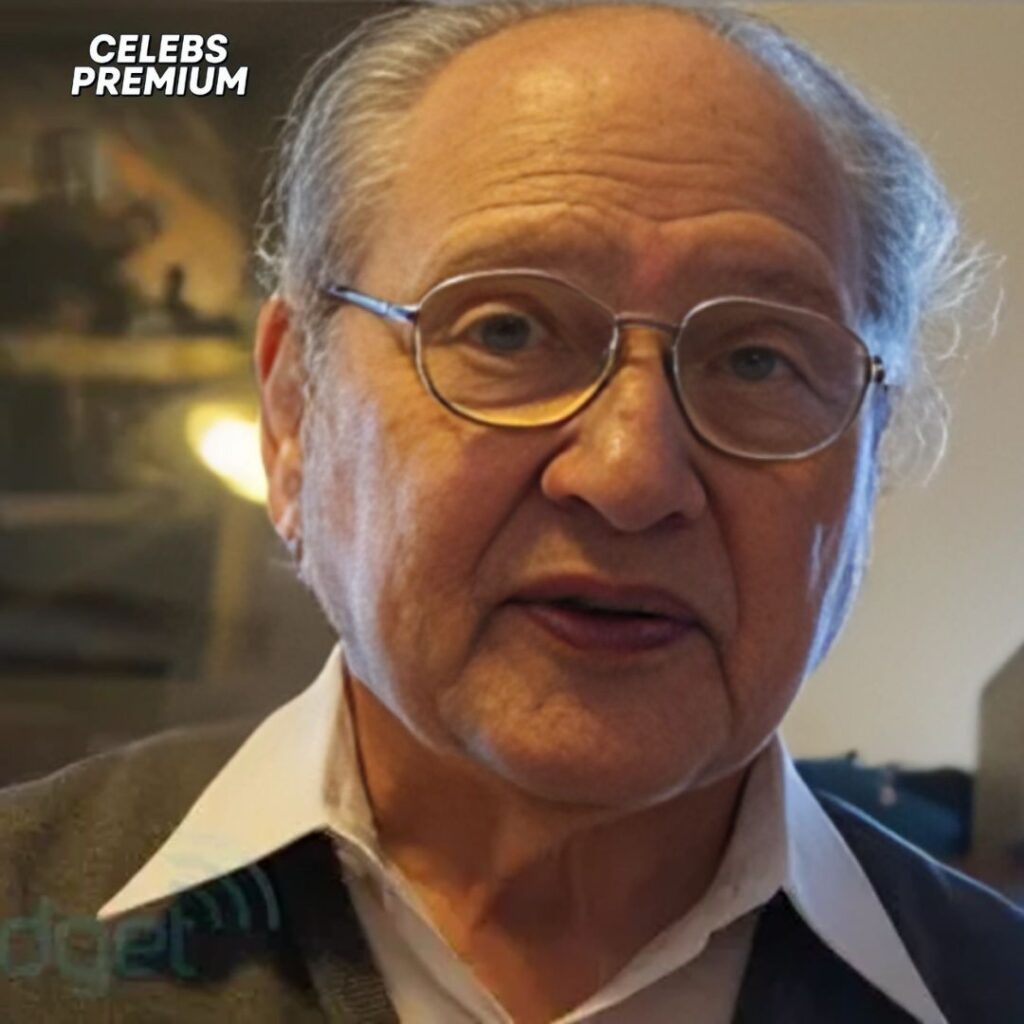 Close-up photo of Ronald Wayne. He is an older man with glasses and a white collared shirt under a dark jacket. He has thinning grey hair and appears to be speaking.